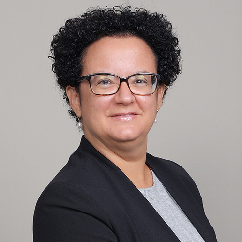 A woman with glasses dressed in a black blazer, smiling softly while standing against a neutral background.