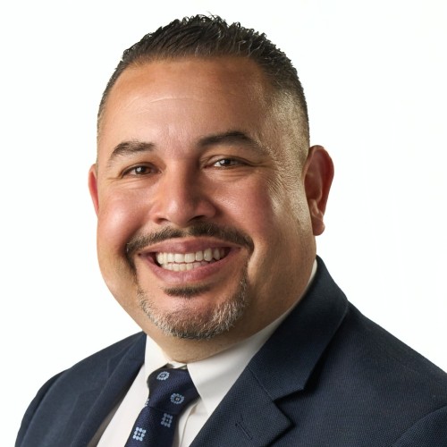 Man in a navy suit, white shirt, and patterned tie, smiling at the camera against a plain white background
