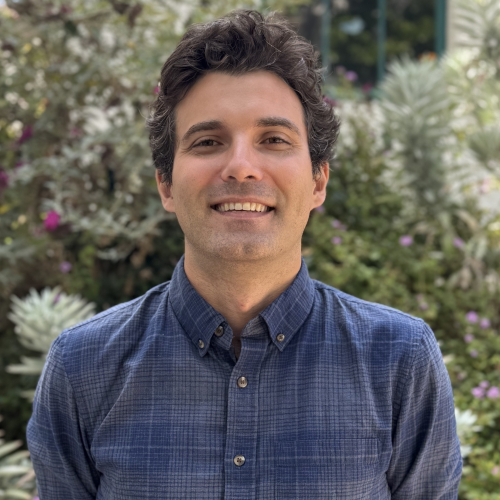A person in a blue checkered shirt smiles standing outdoors, with a background of lush greenery and purple flowers.