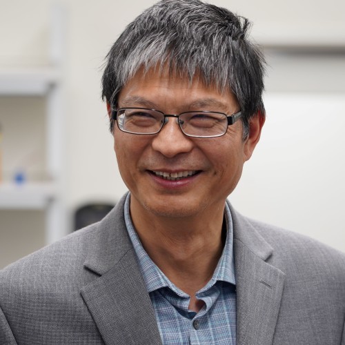 Smiling man in a gray blazer and glasses, standing in a bright indoor setting