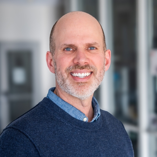 A middle-aged man with a bald head, beard, and blue eyes is smiling at the camera while wearing a blue sweater over a collared shirt.