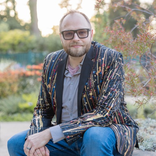 Man in a colorful, artistic blazer sitting in a garden, surrounded by flowers and greenery