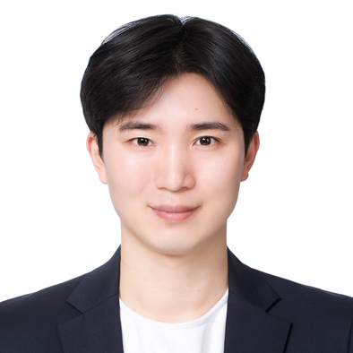 A person with short black hair smiles gently at the camera wearing a dark blazer over a white shirt, set against a plain white background.