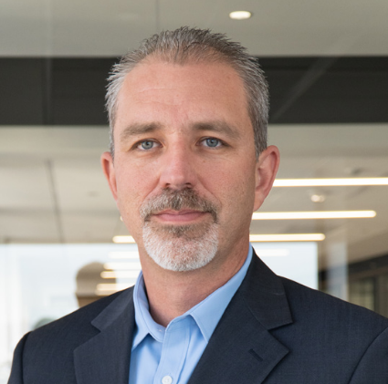 Professional headshot of a man with salt-and-pepper hair and beard, wearing a navy suit and blue shirt, looking calmly at camera.