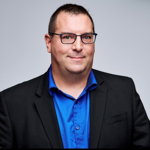 A man wearing glasses, a blue shirt, and a black blazer smiles against a plain grey background.