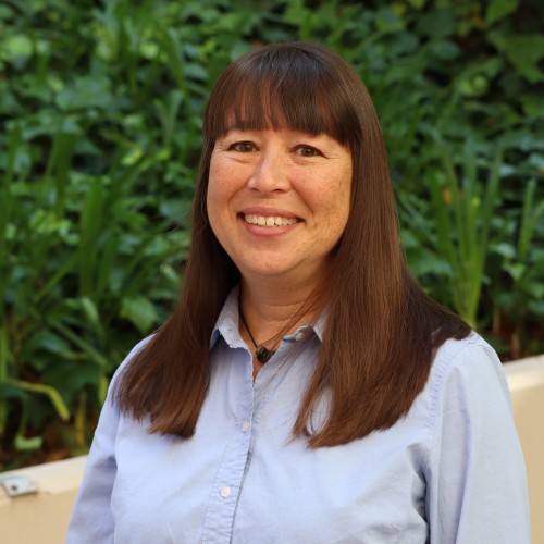 Smiling woman with long brown hair wearing a light blue shirt, standing in a lush green outdoor setting