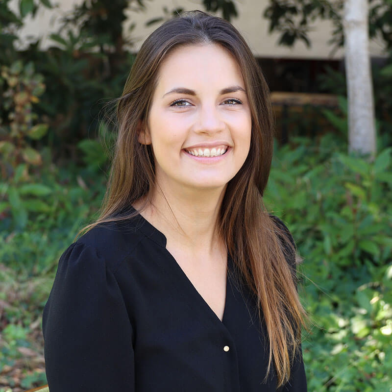 Portrait of a smiling woman with long brown hair wearing a black blouse, posing outdoors amid lush green foliage in warm natural light.