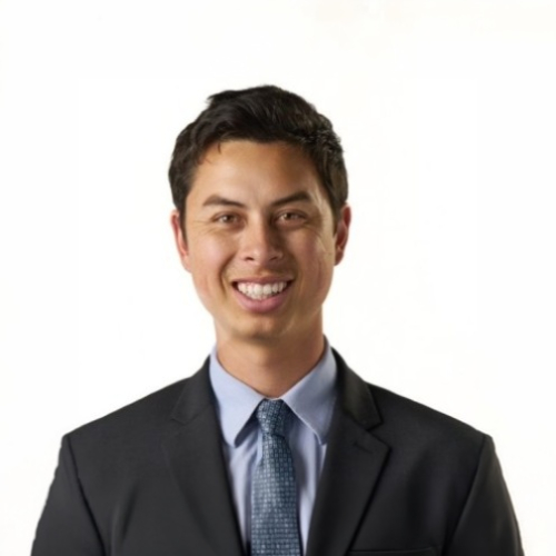Professional head-and-shoulders portrait of a smiling man in a black suit, blue shirt, and patterned tie against a clean white background.