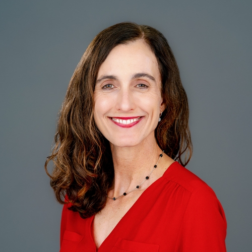 Smiling woman in a red blouse against a gray background. She has shoulder-length wavy hair and wears a simple necklace.