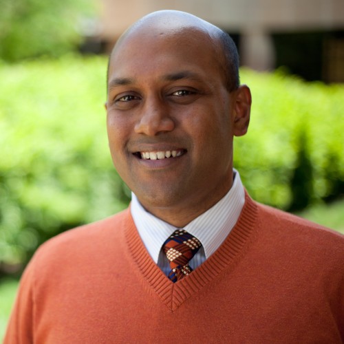 Smiling man in an orange sweater and patterned tie, standing outdoors with greenery in the background